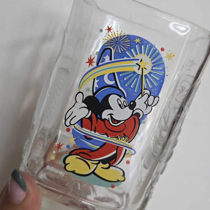 May include: Clear glass tumbler featuring a colorful graphic of Mickey Mouse in a sorcerer's outfit, holding a wand. The design includes a blue background with fireworks and stars. The Disney logo is printed at the bottom.