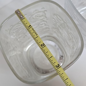 May include: Clear, square-shaped glass with embossed Coca-Cola script. The glass measures approximately 3 inches across. A yellow measuring tape is placed diagonally across the glass, showing the measurement in inches.