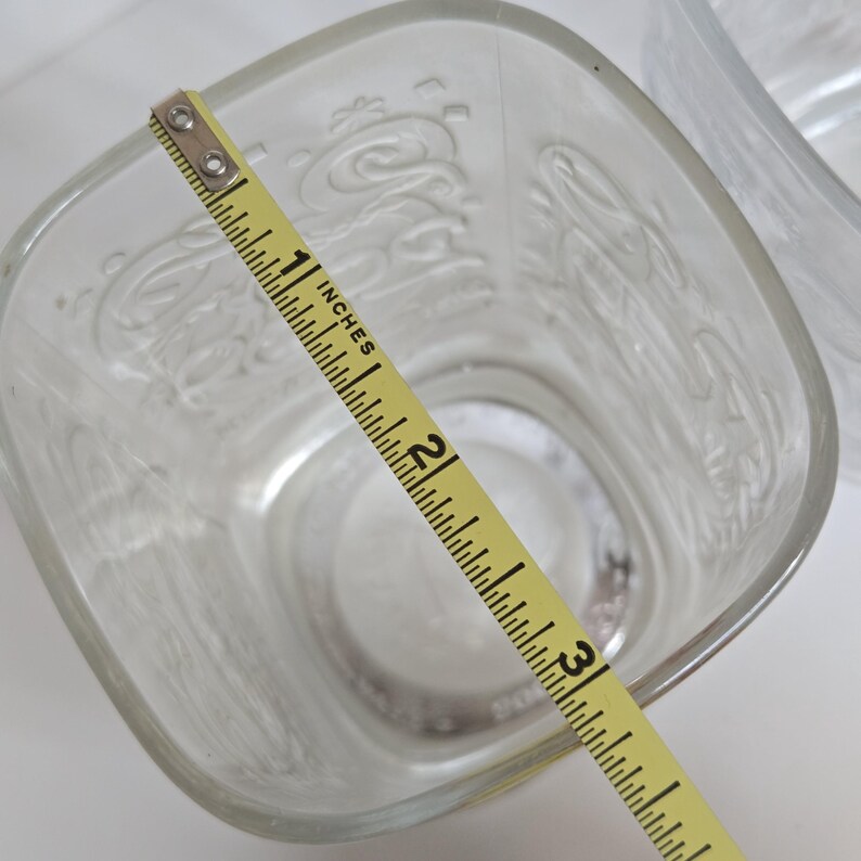 May include: Clear, square-shaped glass with embossed Coca-Cola script. The glass measures approximately 3 inches across. A yellow measuring tape is placed diagonally across the glass, showing the measurement in inches.