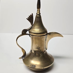 Puede incluir: Una cafetera de latón antigua con un pico largo, un asa curvada y una tapa ornamentada. La cafetera tiene una base bulbosa y un cuello alto y delgado. El latón tiene un tono dorado cálido.