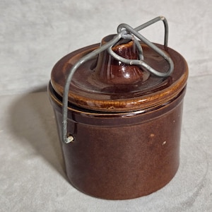 May include: A brown ceramic jar with a lid and a metal handle. The jar is cylindrical with a slightly tapered base. The lid has a raised center and a metal handle that arches over the top. The jar is glazed with a glossy finish.