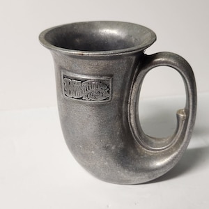 Könnte beinhalten: Ein becherfarbener Becher mit einem einzigartigen, hornförmigen Griff. Der Becher hat ein rechteckiges Emblem mit dem Text "The Swindler" in einer verzierten Schriftart. Der Becher hat einen leicht ausgestellten Rand und ein glattes, mattes Finish.