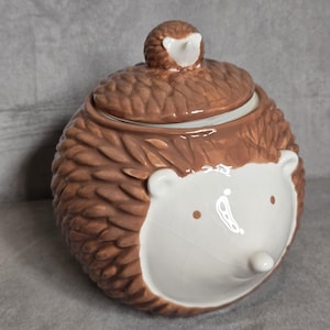 Op de afbeelding: Een keramische pot in de vorm van een egel met een deksel. De pot is voornamelijk bruin met een getextureerd, stekelig ontwerp en een witte gezicht met kleine bruine ogen en een neus. De deksel heeft een kleine egelfiguur.
