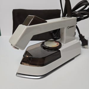 以下が含まれることがあります： 白と茶色のNorelcoトラベルアイロン、黒い電源コード付き。アイロンには茶色の水タンクと温度調節ダイヤルが付いています。背景には茶色のトラベルバッグがあります。