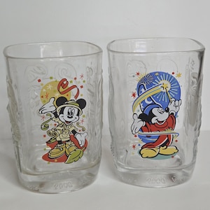 May include: Two clear glass tumblers featuring Mickey Mouse designs. One tumbler shows Mickey in safari attire, the other depicts Mickey as a magician. Both glasses have the year 2000 printed on the base.