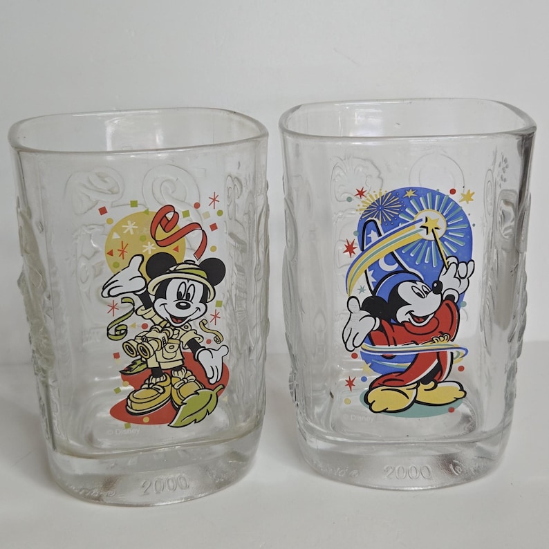 May include: Two clear glass tumblers featuring Mickey Mouse designs. One tumbler shows Mickey in safari attire, the other depicts Mickey as a magician. Both glasses have the year 2000 printed on the base.