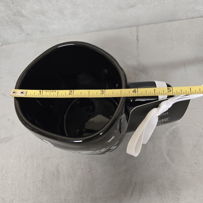 May include: A black ceramic mug with a glossy finish, featuring a white ribbon and a black tag with text. A yellow measuring tape is placed across the top opening, showing a diameter of approximately 5 inches. The mug has a wide, rounded rim.