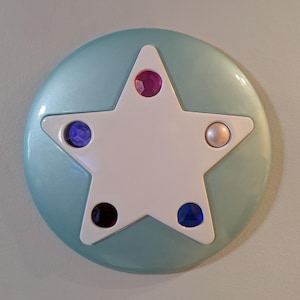 Op de afbeelding: Een cirkelvormig lichtblauw object met een grote witte ster in het midden. De ster is versierd met vijf ingebedde edelstenen: een roze bovenaan, een paarse links, een donkere linksonder, een blauwe rechtsonder en een bleke parelachtige bol aan de rechterkant.