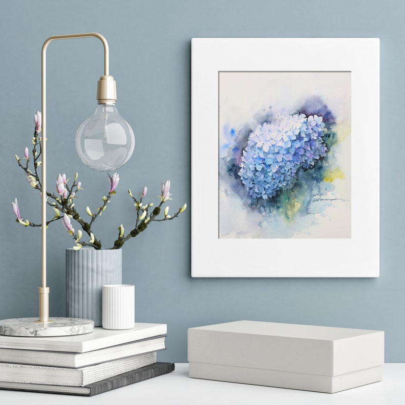 May include: A watercolor painting of a blue hydrangea flower in a white frame. The painting is on a white wall with a gold lamp and a vase of white flowers.
