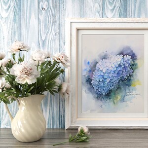 May include: A watercolor painting of a blue hydrangea flower in a white frame. The painting is on a wooden background with a bouquet of white flowers in a cream pitcher.