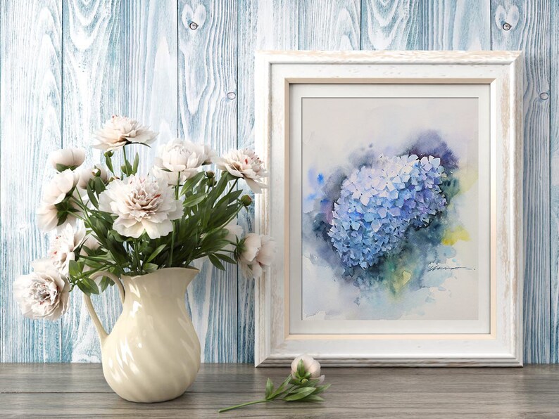 May include: A watercolor painting of a blue hydrangea flower in a white frame. The painting is on a wooden background with a bouquet of white flowers in a cream pitcher.