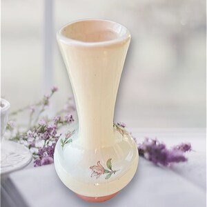May include: A cream-colored ceramic vase with a flared top and a rounded base. The vase features delicate pink and green floral designs. The base is a soft coral color. The vase is likely used for displaying flowers.