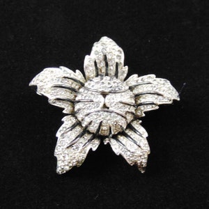 May include: A silver flower brooch with a center made of rhinestones. The flower has five petals and is set against a black background.