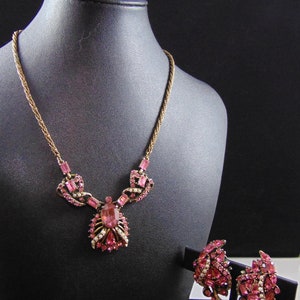 May include: A pink rhinestone necklace, earrings, and brooch set. The necklace features a large pink rhinestone pendant with a gold chain. The earrings are clip-on and feature a cluster of pink rhinestones. The brooch is a floral design with pink rhinestones.