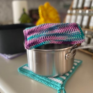 May include: A silver pot with a teal and purple crocheted pot holder on a white countertop.