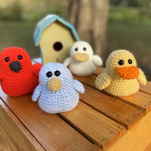May include: Five crocheted stuffed animals, including a red cardinal, a blue bird, a white duck, and two yellow ducks, are arranged on a wooden crate.
