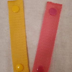 May include: Two fabric straps with plastic buttons. One strap is yellow with yellow buttons and the other is pink with pink buttons.