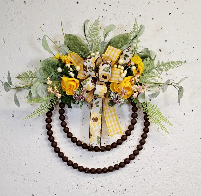 May include: A brown wooden bead wreath decorated with yellow and green floral accents. The wreath features a yellow and white gingham bow with a sunflower design.