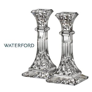 May include: Two clear crystal candlesticks with diamond-cut patterns and square tops. The brand name "WATERFORD" is printed in a dark teal color on the left side. The candlesticks have a classic, elegant design.