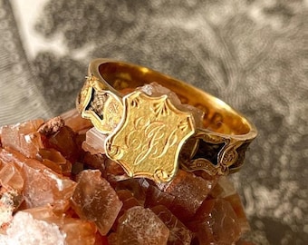 May include: A gold ring with a black enamel band and a gold shield-shaped center with engraved initials. The ring is resting on a cluster of brown crystals.