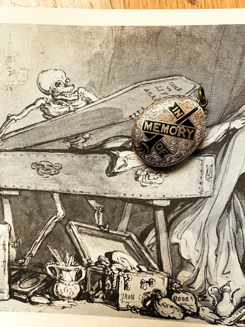 May include: A gold locket with an engraved design and black enamel. The locket has a cross with the words "In Memory Of" on it. The locket is sitting on top of a vintage illustration of a skeleton in a coffin.