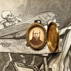 May include: A gold locket with a portrait of a man with a beard inside. The locket is open and the back is visible. The locket is sitting on a piece of paper with a drawing of a skeleton in a coffin. The drawing includes the text "Come to Life Again."