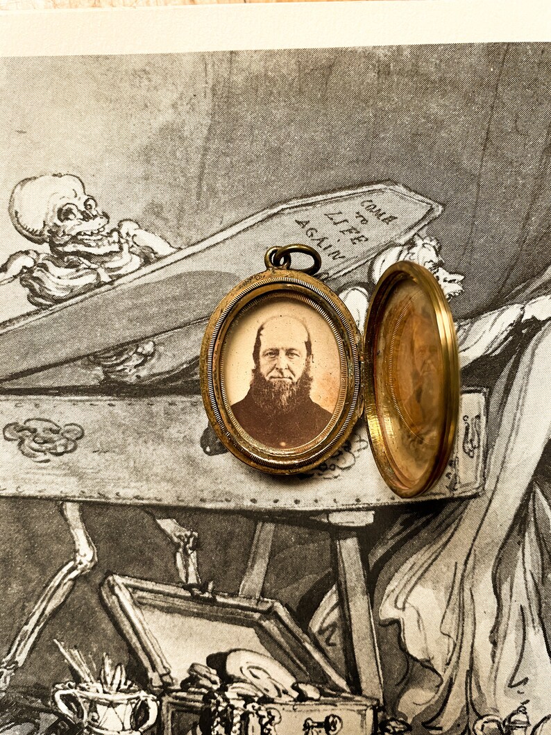 May include: A gold locket with a portrait of a man with a beard inside. The locket is open and the back is visible. The locket is sitting on a piece of paper with a drawing of a skeleton in a coffin. The drawing includes the text "Come to Life Again."