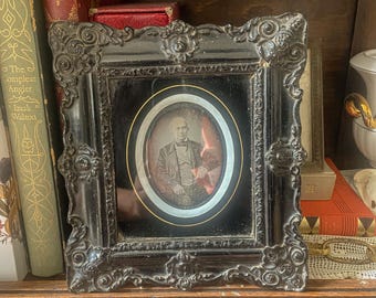 May include: An antique black framed portrait of a man in a suit and tie. The portrait is oval-shaped with a gold border. The frame has ornate detailing and is displayed on a wooden shelf with books.