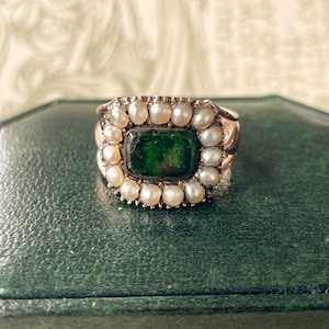 May include: A vintage ring with a rectangular, dark green stone at the center, surrounded by small, round pearls. The ring's band is a gold color, and the pearls are a creamy white. The ring is set on a dark green surface.