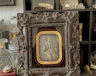 May include: An antique framed portrait with a dark brown, ornate frame. The portrait features a woman in a dress, set against a gold and burgundy background. The frame is highly detailed with floral and scrollwork designs.