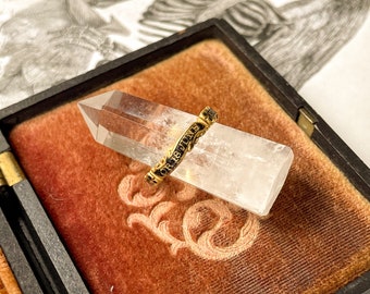 May include: A gold ring with black lettering that reads "OR 18 JUNE" is displayed on a clear crystal. The crystal is resting on a red velvet surface inside a black box.