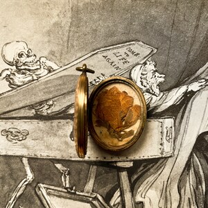 May include: A gold locket containing a pressed flower. The locket is open, revealing the flower. The locket is placed on top of a vintage print depicting a skeleton in a coffin. The print reads "Come to Life Again."