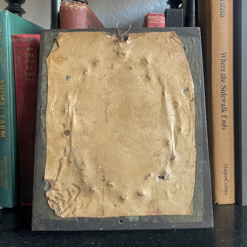 Puede incluir: Una imagen enmarcada vintage con un respaldo de papel marr&oacute;n. El papel tiene varios agujeros y est&aacute; ligeramente rasgado. El marco es negro y tiene un peque&ntilde;o gancho de metal en la parte superior.