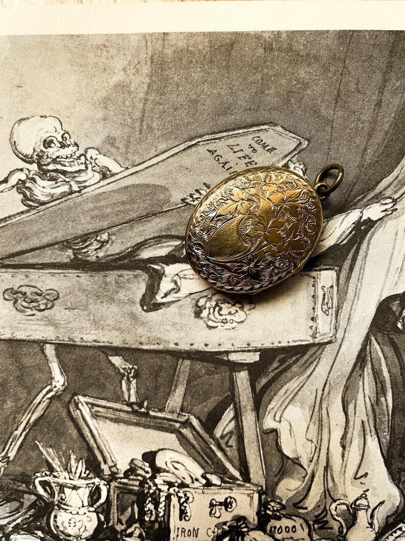 May include: An antique gold locket with an engraved floral design. The locket is resting on a vintage illustration of a skeleton in a coffin. The illustration includes the text "Come to Life Again."