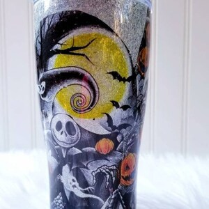 Op de afbeelding: Een zwart-witte tumbler met een glitterafwerking heeft een Nightmare Before Christmas-ontwerp. Het ontwerp bevat Jack Skellington, Sally en andere personages uit de film. De tumbler heeft een rietje en een deksel.