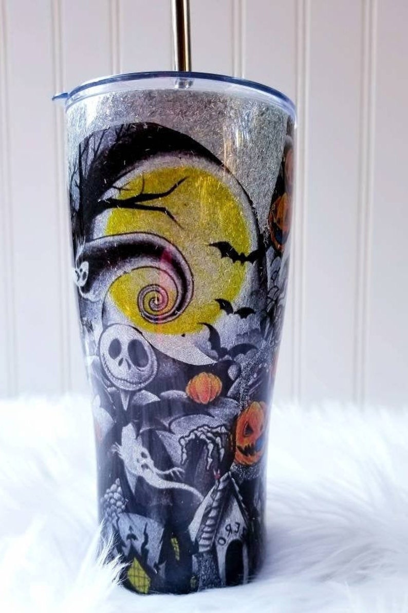 Op de afbeelding: Een zwart-witte tumbler met een glitterafwerking heeft een Nightmare Before Christmas-ontwerp. Het ontwerp bevat Jack Skellington, Sally en andere personages uit de film. De tumbler heeft een rietje en een deksel.