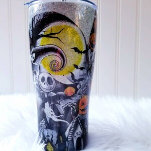 Op de afbeelding: Een zwart-witte tumbler met een glitterafwerking heeft een Nightmare Before Christmas-ontwerp. Het ontwerp bevat Jack Skellington, Sally en andere personages uit de film. De tumbler heeft een zilveren deksel en een doorzichtige rietje.