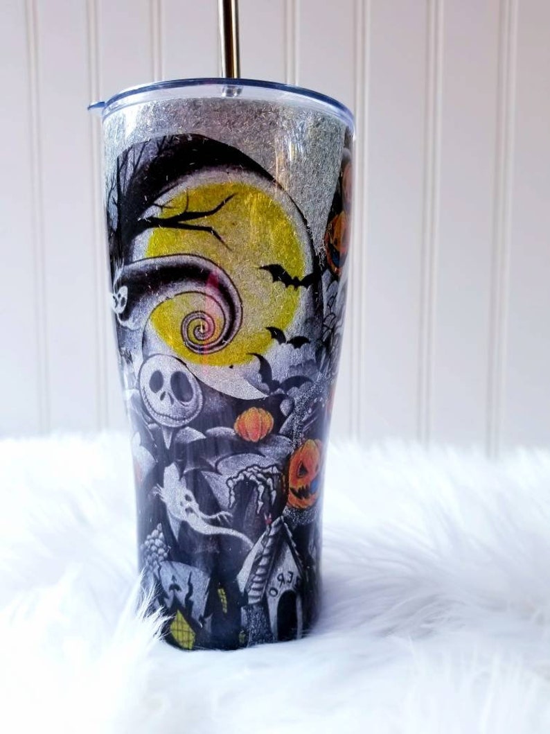 Op de afbeelding: Een zwart-witte tumbler met een glitterafwerking heeft een Nightmare Before Christmas-ontwerp. Het ontwerp bevat Jack Skellington, Sally en andere personages uit de film. De tumbler heeft een zilveren deksel en een doorzichtige rietje.