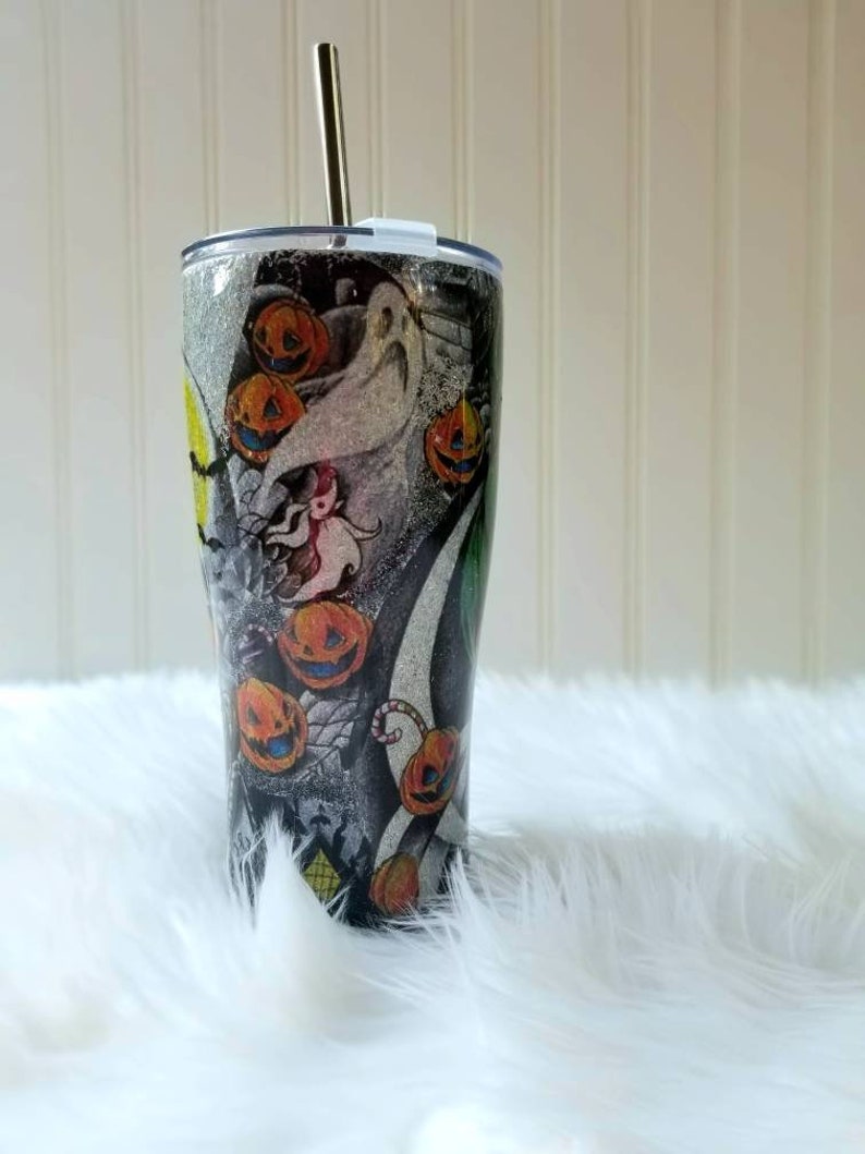 Op de afbeelding: Een zwarte en zilveren roestvrijstalen beker met een Halloween-thema met Jack Skellington en pompoenen. De beker heeft een rietje en een deksel.