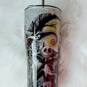 Op de afbeelding: Een zwarte en witte glitterbeker met een Nightmare Before Christmas-ontwerp. Het ontwerp bevat Jack Skellington, Sally en andere personages uit de film. De beker heeft een zilveren deksel en een doorzichtige rietje.