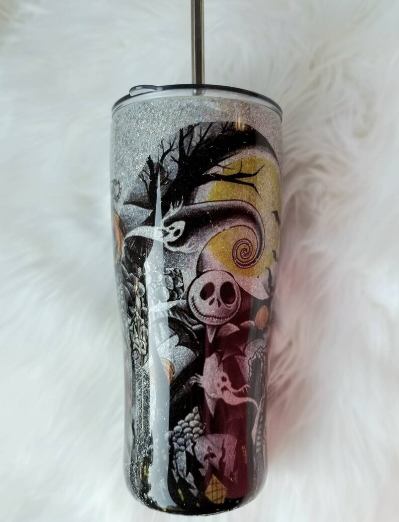 Op de afbeelding: Een zwarte en witte glitterbeker met een Nightmare Before Christmas-ontwerp. Het ontwerp bevat Jack Skellington, Sally en andere personages uit de film. De beker heeft een zilveren deksel en een doorzichtige rietje.