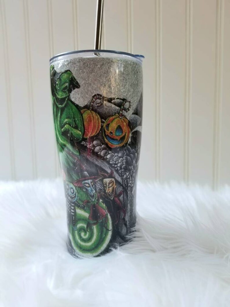 Op de afbeelding: Een zwarte en groene tumbler met een glitterafwerking en een rietje. De tumbler heeft een grafische print met personages uit de film Nightmare Before Christmas, waaronder Jack Skellington, Sally en Oogie Boogie.