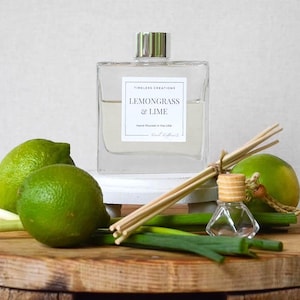 May include: A square glass diffuser bottle with a silver cap and a label that reads "Lemongrass & Lime" sits on a white stand. Two green limes, green onions, and a small glass bottle are on a wooden cutting board.