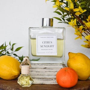 May include: A clear, square glass bottle with a silver cap, labeled "Citrus Sunlight" by Timeless Creations, sits on a wooden surface with lemons, a tangerine, and a small car air freshener. Yellow flowers and greenery add a fresh, natural touch.
