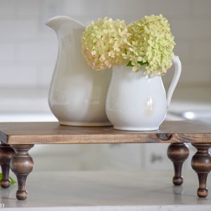 May include: A rustic, wooden riser with turned legs, holding two white ceramic pitchers. One pitcher is filled with light green and pink flowers. The riser is stained a warm brown color, and the background is a blurred kitchen setting.