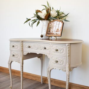 May include: A light beige vanity table with a dark wood top, featuring four curved drawers with ornate floral carvings, slender cabriole legs, and small round metal knobs. A white speckled vase with greenery and a framed picture sit on top.