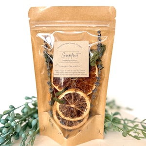 May include: A clear, resealable bag filled with dried grapefruit slices, lavender sprigs, and mint leaves. A small, brown label reads "Grapefruit Simmering Potpourri" with instructions. The bag is set against a white surface with green foliage.