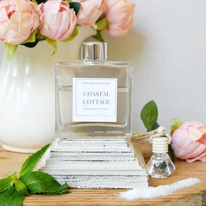 May include: A clear glass bottle with a silver cap and a label that reads "Coastal Cottage" sits on a white, distressed wooden stand. The image includes a white pitcher with pink flowers, a small glass bottle, and a sprig of mint.