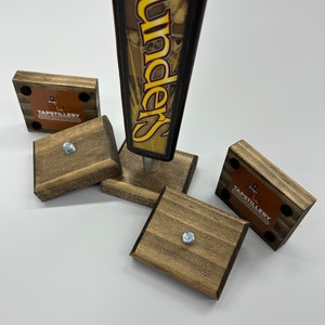 May include: Four wooden tap handles with the text "Tapstillery" and a cross logo. The tap handles are brown and have a distressed finish. The tap handles are arranged in a square shape. A Founders beer tap handle is in the center of the square.