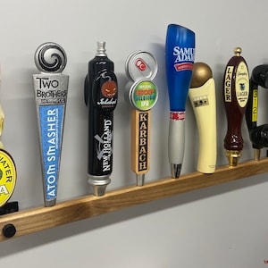 May include: A wooden wall-mounted beer tap handle rack with nine different tap handles. The tap handles feature various designs, including a fish, a pumpkin, a beer can, and a phone. The rack is made of dark wood and has a natural finish. The text "TAP HANDLES NOT INCLUDED" is printed at the bottom of the image.